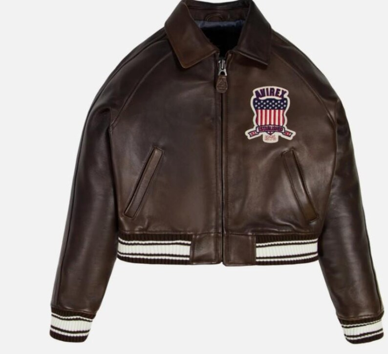May include: A dark brown leather bomber jacket featuring a front zipper closure, a classic collar, and ribbed cuffs and hem with white stripes. An embroidered patch on the chest displays the text AVIREX above a stylized American flag design.