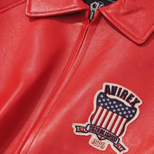 May include: Close-up of a red leather jacket with a collar and zipper. The jacket features a patch with the text "AVIREX" above a design of the American flag and the text "NY ESTABLISHED NY 1975".