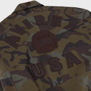 May include: A camouflage jacket featuring a brown, green, and black pattern. The back displays the text AVIREX USA in dark brown lettering, arched above and below a circular emblem, all rendered in a dark brown material against the camo fabric.