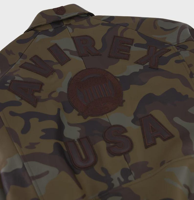 May include: A camouflage jacket featuring a brown, green, and black pattern. The back displays the text AVIREX USA in dark brown lettering, arched above and below a circular emblem, all rendered in a dark brown material against the camo fabric.