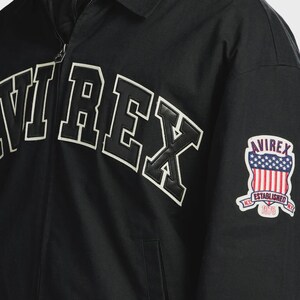 May include: A black zip-up jacket featuring the word AVIREX in large, bold, white-outlined black lettering across the chest. On the sleeve, there is a shield-shaped patch displaying the American flag, the text AVIREX, ESTABLISHED, and 1975.