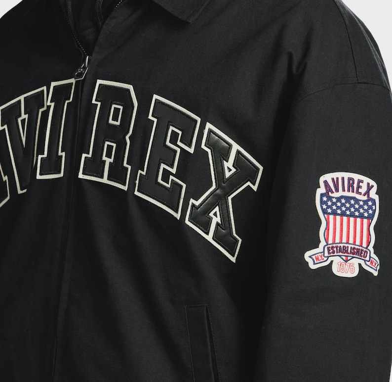 May include: A black zip-up jacket featuring the word AVIREX in large, bold, white-outlined black lettering across the chest. On the sleeve, there is a shield-shaped patch displaying the American flag, the text AVIREX, ESTABLISHED, and 1975.