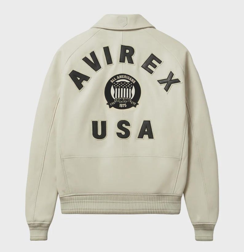 Pode incluir: Casaco de couro creme com gola cl&aacute;ssica e punhos canelados. As costas apresentam a palavra "AVIREX" em letras pretas grandes, juntamente com um emblema circular e a palavra "USA". Um casaco bomber elegante.