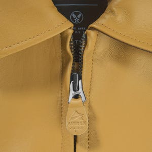May include: Close-up of a mustard-colored leather jacket with a zipper. The zipper is silver and has a leather pull tab with the "AVIREX" logo. The jacket has a collar and a tag with text that reads "CIVILIAN & MILITARY TAILORS".