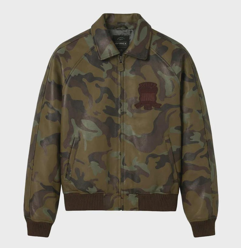 May include: A camouflage print leather jacket featuring a full-length front zipper, a classic collar, and ribbed knit cuffs and hem. The chest displays a dark brown embroidered patch with the text Avirex. The jacket has two side pockets.