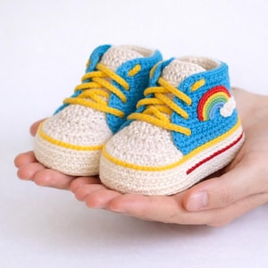 Puede incluir: Un par de zapatillas de bebé de ganchillo hechas a mano. Los zapatos son azules, crema y amarillos, con un arcoíris y una nube aplicados en el lateral. Las zapatillas están sostenidas en las manos de una persona.