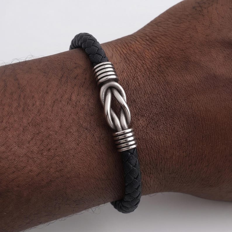 Peut inclure: Bracelet en cuir tress&eacute; noir avec un motif de n&oelig;ud argent&eacute;. Le bracelet pr&eacute;sente des accents argent&eacute;s de chaque c&ocirc;t&eacute; du n&oelig;ud. Le bracelet est port&eacute; au poignet.