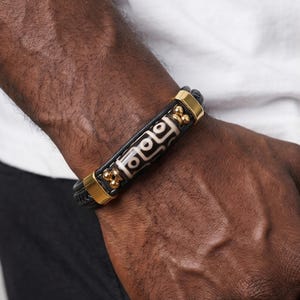 Peut inclure: Bracelet en cuir noir avec une perle rectangulaire centrale ornée d'un motif géométrique blanc et noir. Le bracelet est orné d'éléments métalliques dorés et est porté au poignet.