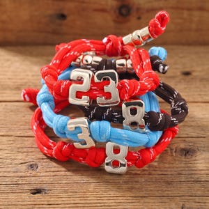 Può includere: Un set di tre braccialetti in corda regolabili nei colori rosso, azzurro e marrone scuro. Ogni braccialetto presenta un ciondolo in metallo argentato a forma di numero, inclusi i numeri 2, 3 e 8, integrati nel design del cordino intrecciato.