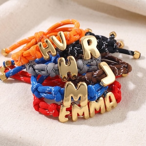 Könnte beinhalten: Eine Sammlung farbenfroher Seilarmbänder mit goldfarbenen Buchstabenanhängern, die Namen und Initialen buchstabieren. Die Armbänder haben orange, schwarze, graue, braune, rote und blaue Seile mit goldenen Akzenten. Die Buchstaben sind fett und ohne Serifen.