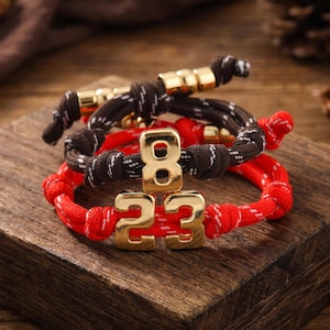 May include: Three bracelets with gold-colored numbers and rope-like bands. The top bracelet is brown and white, the middle one is red and white, and the bottom one is red and white. The numbers are "8" and "23".