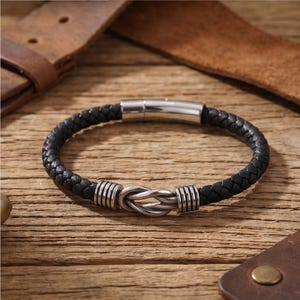 Peut inclure: Bracelet en cuir tress&eacute; noir avec fermoir et n&oelig;ud en acier inoxydable. Le bracelet est pr&eacute;sent&eacute; sur une surface en bois avec des accents de cuir. Un accessoire &eacute;l&eacute;gant.
