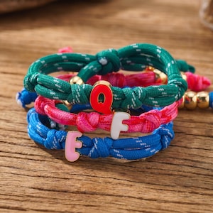 Puede incluir: Pila de tres pulseras coloridas con las letras "E", "F" y "Q". Las pulseras están hechas de cuerda anudada en verde, rosa y azul. Cada pulsera tiene un acento dorado y un dije de letra.