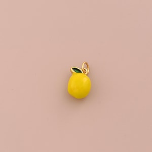 May include: A small, bright yellow lemon-shaped charm with a smooth, glossy enamel finish. The charm features a single, small green leaf detail at the top, attached to a gold-toned metal jump ring for hanging on a necklace or bracelet chain.