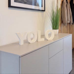 Könnte beinhalten: Vier einzelne weiße, dreidimensionale Blockbuchstaben, die das Wort YOLO bilden. Jeder Buchstabe ist freistehend mit einer glatten, matten Oberfläche und in einer horizontalen Linie auf einer flachen Oberfläche angeordnet, was ein klares Display ergibt.