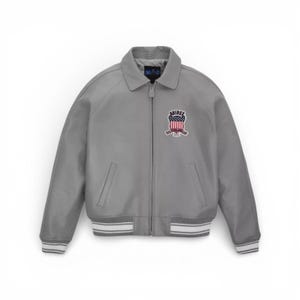 Puede incluir: Chaqueta bomber gris con cuello y cierre de cremallera. La chaqueta presenta un parche con un dise&ntilde;o rojo, blanco y azul y la palabra "AMIREX". Los pu&ntilde;os y el dobladillo tienen detalles a rayas blancas y grises.