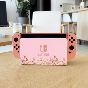 Könnte beinhalten: Eine Nintendo Switch-Konsole mit einem rosa Blumendesign. Die Konsole hat korallenfarbene Controller und ein rosa Dock mit dem Nintendo Switch-Logo. Das Dock hat ein Blumenmuster am unteren Rand.