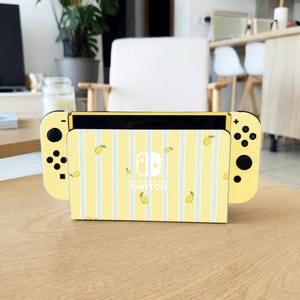 Peut inclure: Une console Nintendo Switch jaune avec des manettes assorties. La station d'accueil de la console présente un motif à rayures jaunes et blanches avec des illustrations de citrons. Le logo Nintendo Switch est visible.