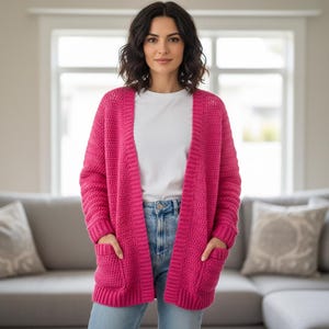 Könnte beinhalten: Ein leuchtend pinkfarbener Cardigan mit offener Front und zwei Vordertaschen. Der grob gestrickte Pullover wird über einem weißen T-Shirt und einer blauen Jeans getragen. Der Cardigan hat eine kräftige, einfarbige Farbe und scheint aus einem weichen, warmen Material zu sein.