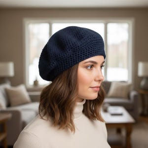 May include: A navy blue knit beret. The beret is made of a textured knit fabric and is worn on the head. The person in the image is wearing a cream-colored turtleneck sweater. The background is a blurred interior scene.