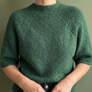May include: A forest green knit sweater featuring a ribbed crew neckline and short sleeves. The garment is constructed with a visible raglan sleeve seam detail and a textured, speckled yarn finish throughout the entire knitted fabric surface.