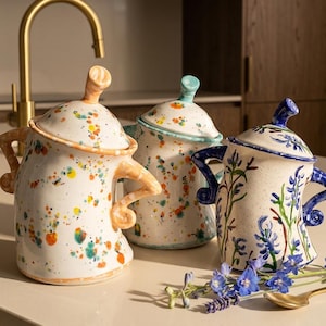 May include: Three ceramic canisters with lids and curved handles. Two canisters feature a multicoloured paint splatter pattern in orange, yellow, and teal. The third canister displays a hand-painted blue floral design on a cream-coloured base.