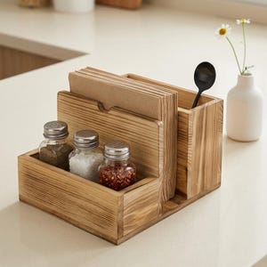 Op de afbeelding: Een houten keukenorganizer met drie glazen kruidenpotjes, een sectie voor servetten en een gleuf voor een zwarte lepel. De potjes bevatten peper, zout en rode pepervlokken. Een kleine witte vaas met bloemen staat op de achtergrond.