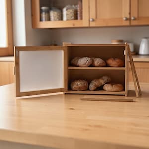 Puede incluir: Una panera de madera de dos niveles con una puerta transparente, abierta hacia un lado. La panera contiene varias hogazas de pan. La panera es de madera clara y está sobre una superficie de madera.