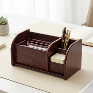Puede incluir: Un organizador de escritorio de madera marrón oscuro con múltiples compartimentos. Cuenta con una sección para bolígrafos, una ranura para notas adhesivas y un área central para objetos pequeños. El organizador está sobre un escritorio claro.