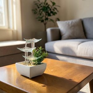 以下が含まれることがあります： 段になった透明ガラスのデザインの、小さな屋内用噴水。水は、小さな緑色の植物が入った白い正方形のベースに、段を流れ落ちます。噴水は、リビングルームの木製のテーブルの上にあります。