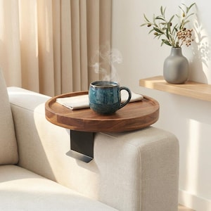 May include: A round, dark wood sofa arm tray with a black metal bracket. A blue ceramic mug with steam rising from it sits on the tray, along with a white folded cloth. A neutral-toned sofa is in the foreground.