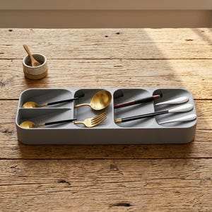 Puede incluir: Un organizador de cubiertos de pl&aacute;stico gris con m&uacute;ltiples compartimentos, que contiene varios utensilios dorados y negros. Los utensilios incluyen cucharas, tenedores y cucharas de servir. Tambi&eacute;n hay una peque&ntilde;a taza de cer&aacute;mica con una cuchara de madera.