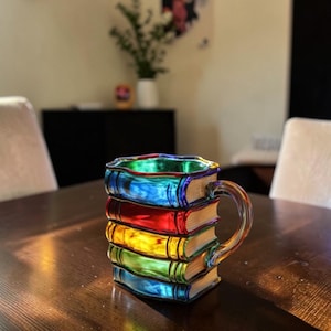 Puede incluir: Una taza de vidrio de colores en forma de pila de libros. La taza presenta un arcoíris de colores, incluyendo azul, rojo, amarillo y verde. El asa es de vidrio transparente. La taza está sobre una mesa de madera.