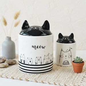 May include: Two white ceramic cat-themed canisters with black accents. The larger canister has a black lid shaped like a cat's head and the word "meow". The smaller canister also has a cat head lid. Both have cat illustrations.