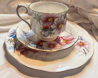 Puede incluir: Juego de té vintage de porcelana blanca. El conjunto incluye una taza, un platillo y un plato, decorados con flores rosas, rojas, azules y amarillas. Los bordes son dorados. El conjunto está sobre una superficie blanca.
