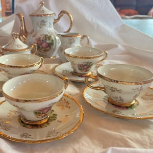 Puede incluir: Un juego de té de porcelana vintage con ribete dorado. El juego incluye una tetera, azucarero, lechera y varias tazas y platillos. Cada pieza presenta un diseño floral con detalles en rosa y dorado, creando una estética elegante y clásica.