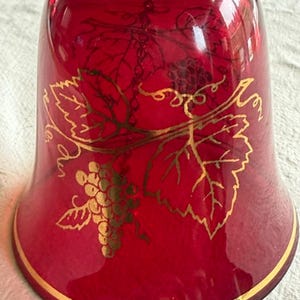 Könnte beinhalten: Eine rubinrote Glasglocke mit einem goldfarbenen Motiv aus Weintrauben und Weinblättern. Die Glocke hat eine glatte, durchscheinende Oberfläche mit einem dünnen Goldrand am unteren Ende und einem gewellten Rand am oberen Griffbereich.