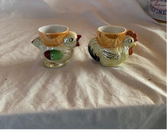 Puede incluir: Dos hueveras de cerámica con forma de gallinas. Una es un pollito amarillo con detalles rojos y verdes, y la otra es un gallo con detalles negros, amarillos y rojos. Las hueveras tienen un soporte dorado para la taza.