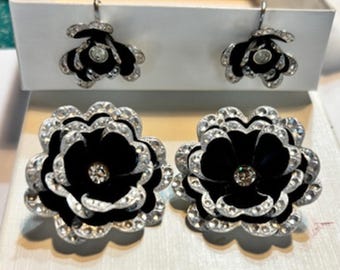 Puede incluir: Un conjunto de pendientes y pendientes a juego con forma de flor negra y plateada. Los pendientes tienen un centro negro con pétalos plateados y un cristal transparente en el centro. Los pendientes se muestran en una caja blanca.