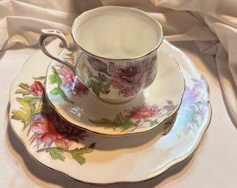Puede incluir: Un juego de taza de té, platillo y plato vintage. Las piezas de porcelana blanca presentan un diseño floral en tonos de rosa, morado y verde, con ribetes dorados. La taza de té se encuentra sobre el platillo, que descansa sobre un plato más grande.