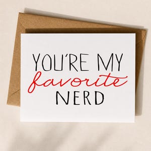 May include: A white greeting card with the text "YOU'RE MY favorite NERD" in black and red lettering. The card is placed on top of a brown envelope. The card is a square shape.