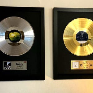 Pode incluir: Dois prêmios de disco emoldurados. Um é prateado com uma imagem de maçã verde, o outro é dourado. Ambos estão em molduras pretas com placas abaixo. O disco de prata é para The Beatles, Abbey Road.