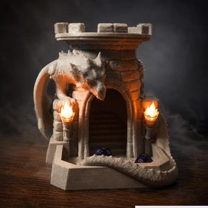 May include: A light-coloured stone-textured dice tower shaped like a castle turret with a dragon perched on the side. Two small orange LED torches flank the arched entrance, and two dark blue polyhedral dice sit at the base. Text reads Physical Version.
