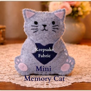 Könnte beinhalten: Eine graue Filzkatze mit rosa Ohren und Pfoten, die ein marineblaues Herz hält. Der Text "Keepsake Fabric" und "Mini Memory Cat" ist auf dem Körper der Katze abgebildet. Die Katze sitzt auf einer Spitzefläche.