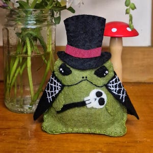 May include: A green felt frog plushie wearing a black top hat with a red band and a black cape featuring white spiderweb embroidery. The frog holds a small white skull in its hand, with black stitched eyes and a simple mouth line on its face.