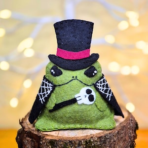 May include: A green felt frog plushie wearing a black top hat with a magenta band. The frog has black cape-like wings with white spiderweb embroidery and holds a small white skull on a black stick. The stitching is visible along the edges.