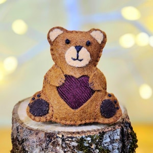 Op de afbeelding: Een bruine vilten teddybeer met een donkerpaarse hartvormige applicatie op de borst, crèmekleurige binnenoren en snuit, en donkerbruine stiksels voor de ogen, neus, mond en voetzooltjes, afgewerkt met zichtbare festonsteken.