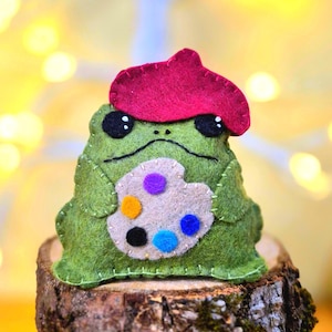 Peut inclure: Une peluche en feutrine verte en forme de grenouille portant un béret rose vif. La grenouille a des yeux noirs avec des reflets blancs et tient une palette d'artiste en feutrine beige avec des pastilles en violet, orange, noir et bleu.