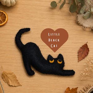 May include: A black felt cat with yellow eyes stretches out on a wooden surface. A brown heart-shaped graphic with the words "LITTLE BLACK CAT" is above the cat. Autumn leaves and decorative elements surround the cat.