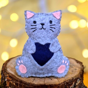 May include: A grey felt cat ornament featuring pink inner ears and paw pads, with black embroidered eyes, nose, and whiskers. The cat holds a dark blue fabric heart centred on its chest, with visible stitching along the edges of the felt body.